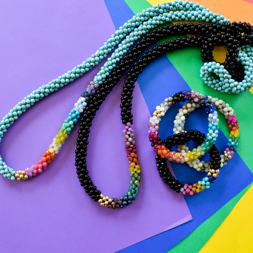 rainbow, black onyx and gold beaded necklace with other rainbow gemstone necklaces and bracelets on colorful background